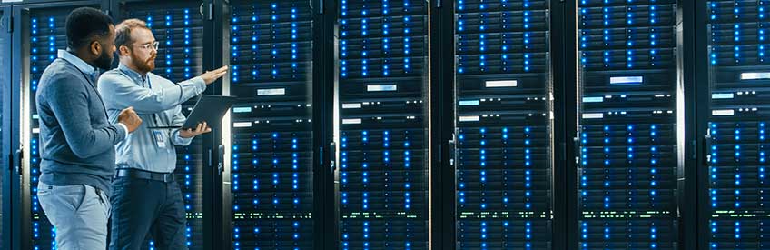 IT Technician with a Laptop Computer and Black Male Engineer Colleague are Talking in Data Center while Walking Next to Server Racks.