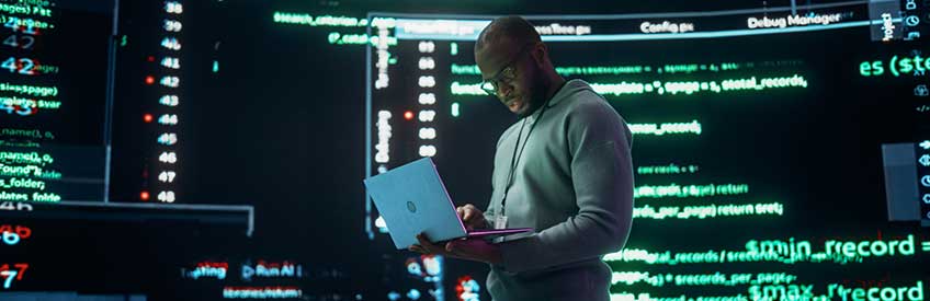 Portrait of Young Black Man Working on Laptop Computer, Looking at Big Digital Screens Glitching While Displaying Code Lines.